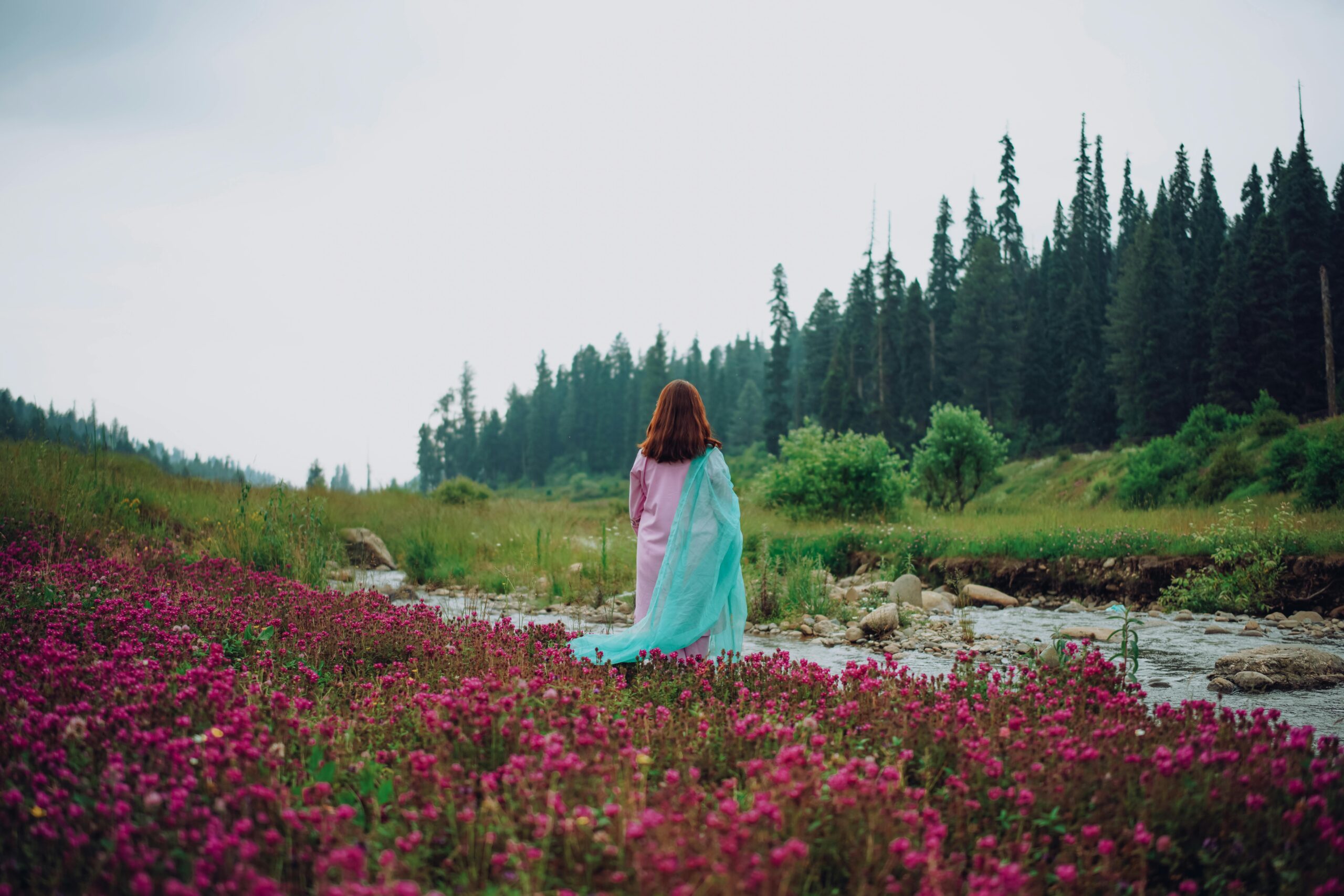 A serene scene of a woman standing in a meadow, adorned with a pink dress and blue veil, next to a flowing stream.